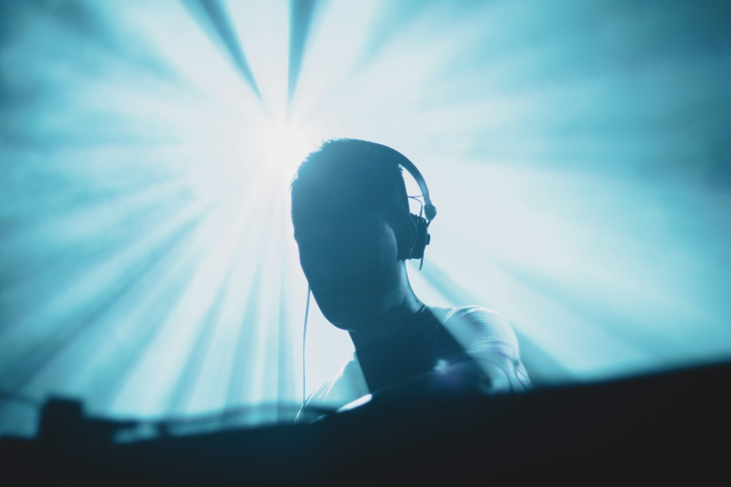 Silhouette einer Person mit Kopfhörern, die vor einer hellblauen Bühnenbeleuchtung steht, deren Strahlen nach außen gerichtet sind.
